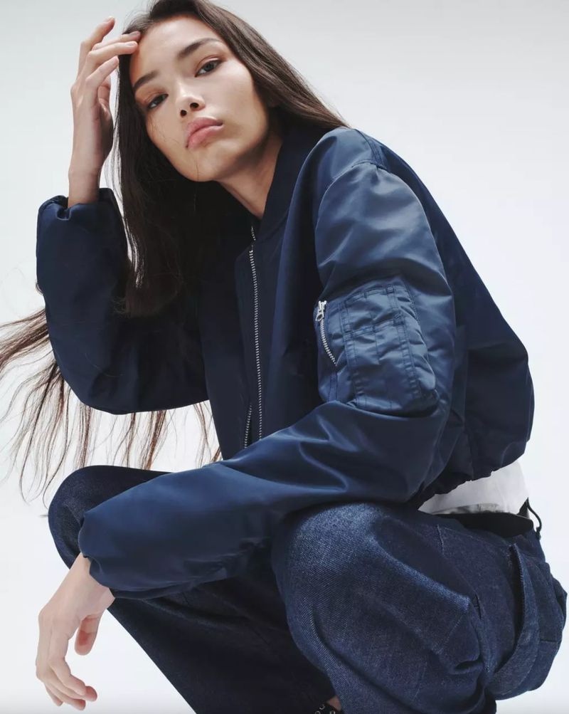 Young woman in a navy bomber jacket and jeans squats in a studio, looking at the camera in a fashion pose with hand in her hair.