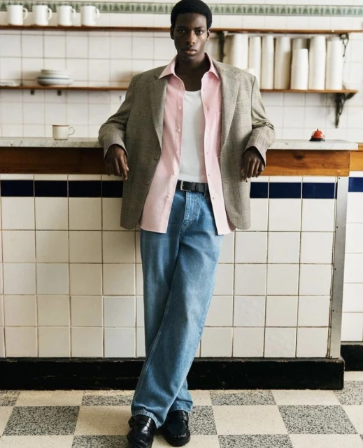 Stylish African-American man in an unbuttoned pink shirt, oversized blazer, and jeans poses indoors near tiled walls and shelves.