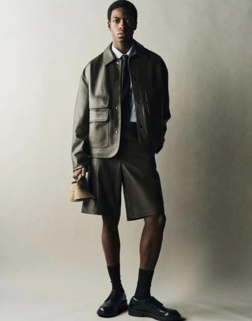 Full studio shot of a dark-skinned adult male wearing a leather jacket and shorts with black shoes, socks, and holding a shoe.