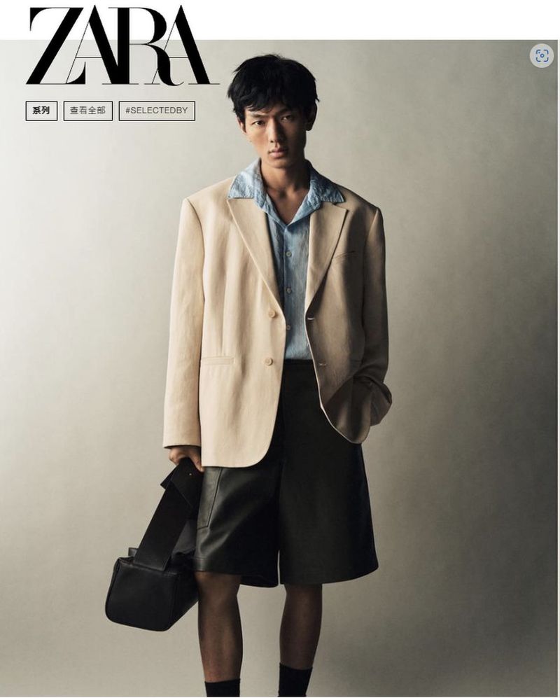 A stylish Asian male model poses in a beige blazer, blue shirt, and dark shorts, holding a black bag against a neutral backdrop.