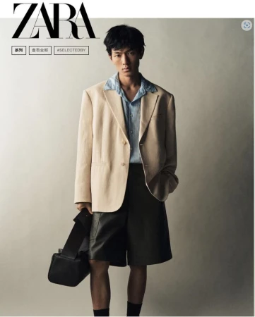 A stylish Asian male model poses in a beige blazer, blue shirt, and dark shorts, holding a black bag against a neutral backdrop.