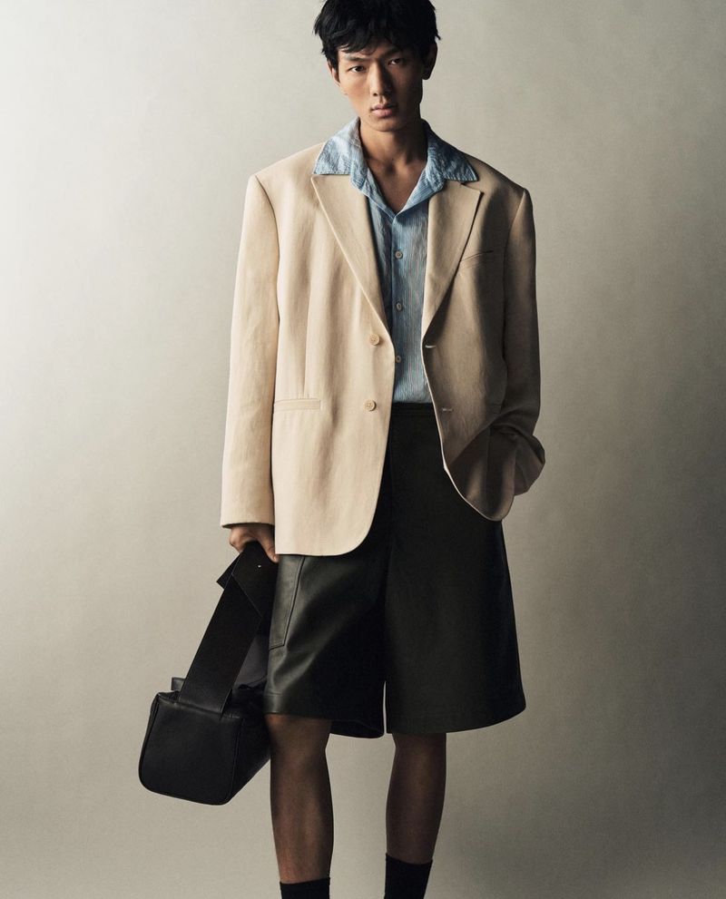 Portrait of a young Asian man in a beige blazer and dark shorts carrying a black bag, posing against a neutral background in a studio setting.