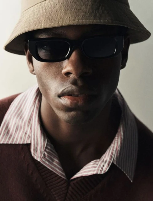 Close-up of a stylish Black man wearing sunglasses and a tan bucket hat against a neutral backdrop, fashion portrait.