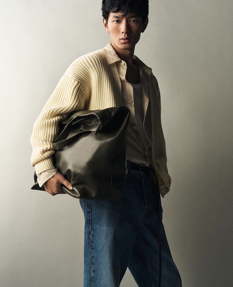 Asian man poses with a large olive-green leather tote bag, showcasing a casual, fashion-forward look with jeans and a knit cardigan.