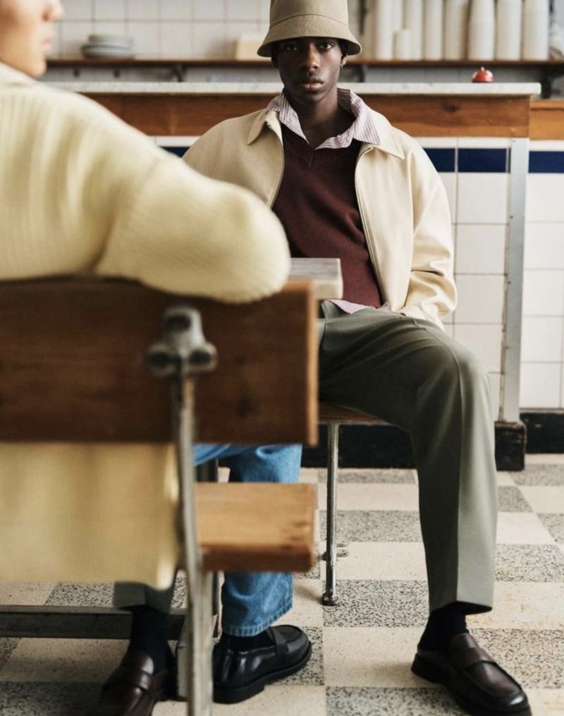 Two people sit at a table indoors, one wears a bucket hat and jacket, the other wears a cream sweater, in a casual setting.