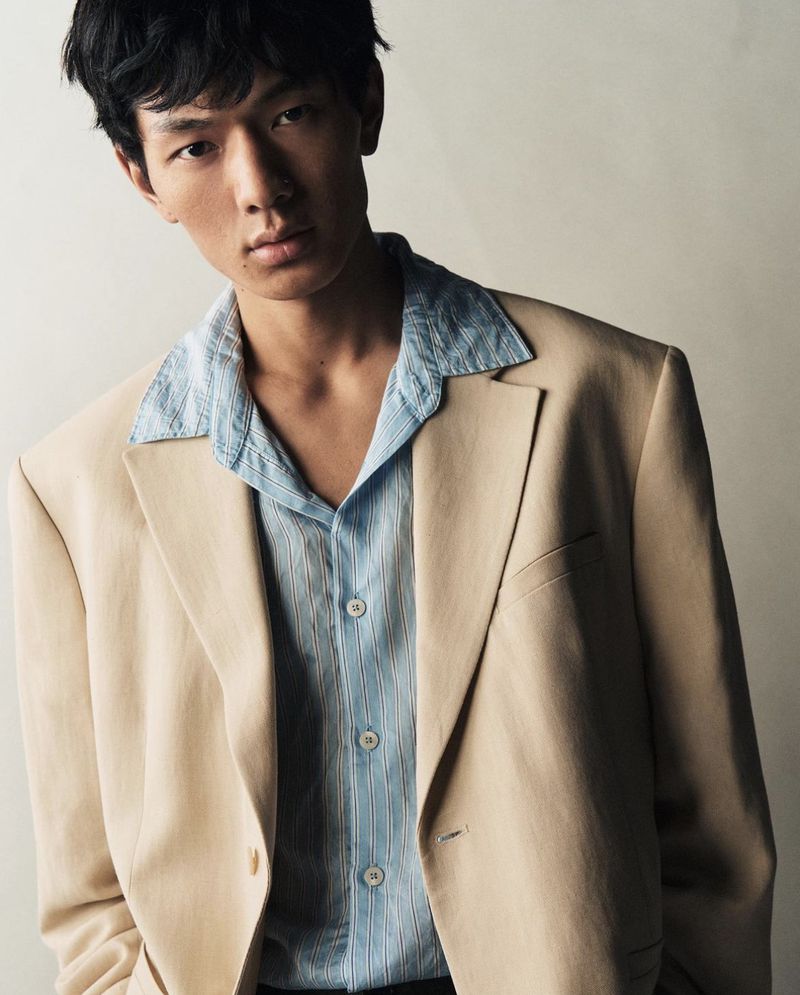 Headshot of a young Asian man wearing a tan blazer and light blue button-down shirt, looking directly at the camera in a studio.