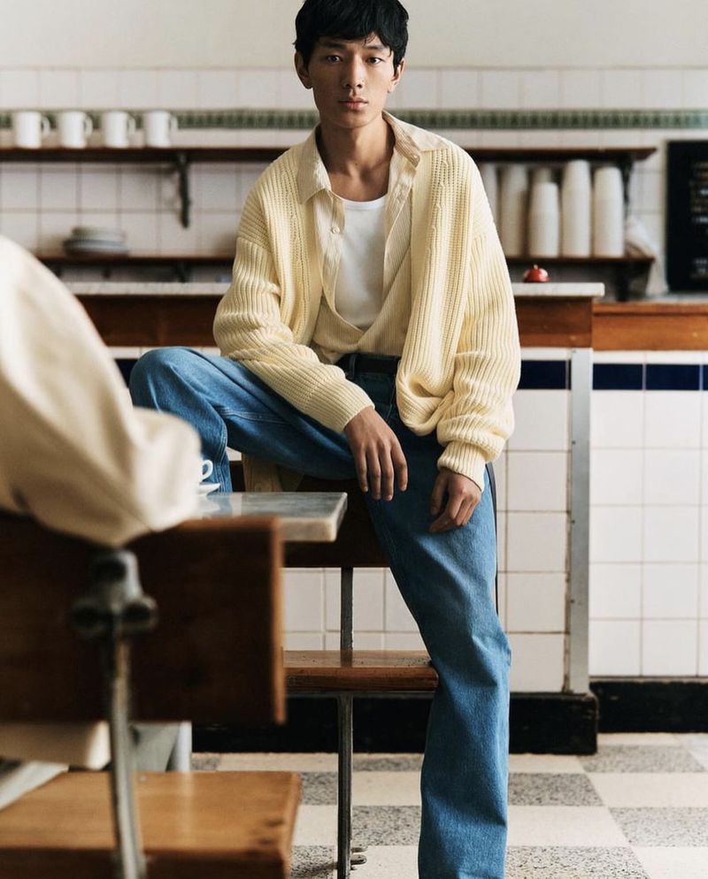 A casually dressed young man in a yellow sweater and blue jeans sits on a table in a sunlit room with a tile floor and neutral walls.