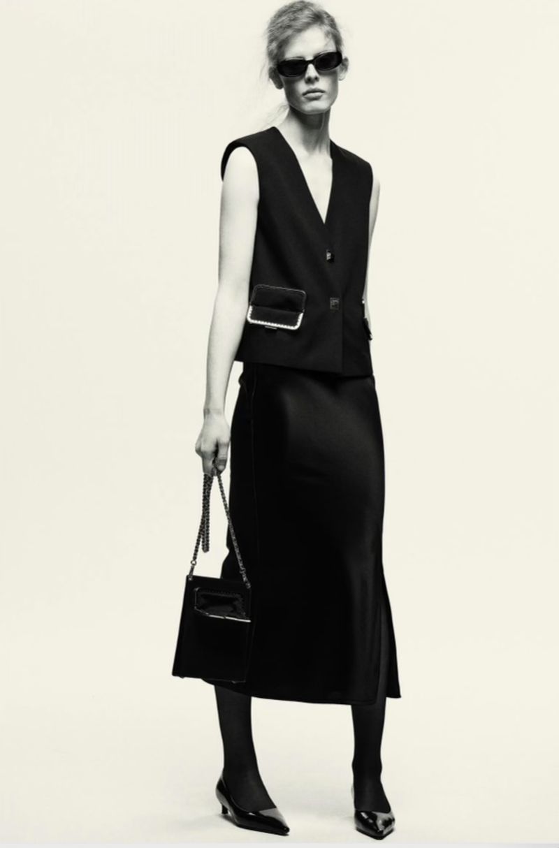 Elegant adult female wearing a black skirt and vest holding a black purse in a monochrome studio shot with clean white backdrop.