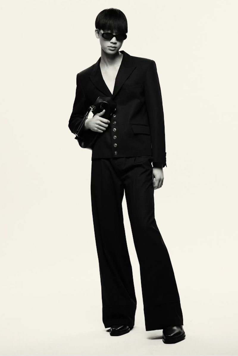 Model wearing a black tailored blazer, wide-leg trousers, and dark sunglasses holding a clutch against a pale studio backdrop.
