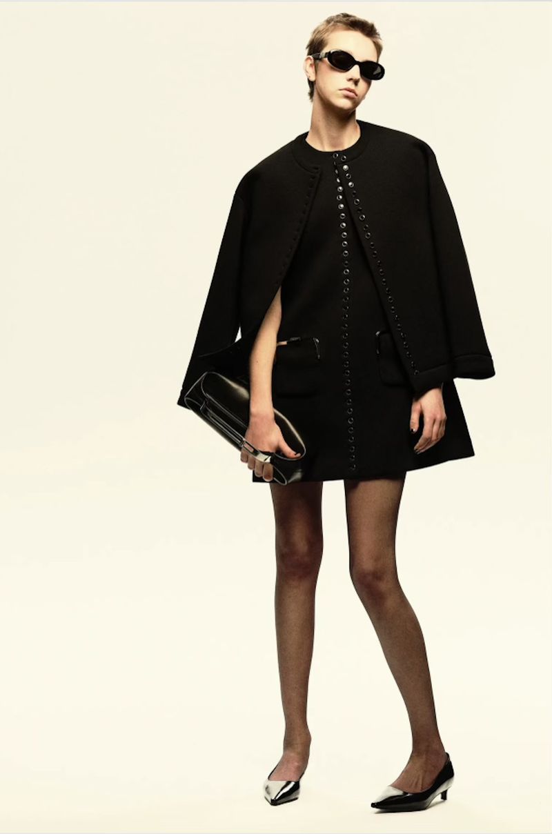 Fashionable woman posing in a black dress, cape, sunglasses, and heels against a cream backdrop, holding a black clutch.