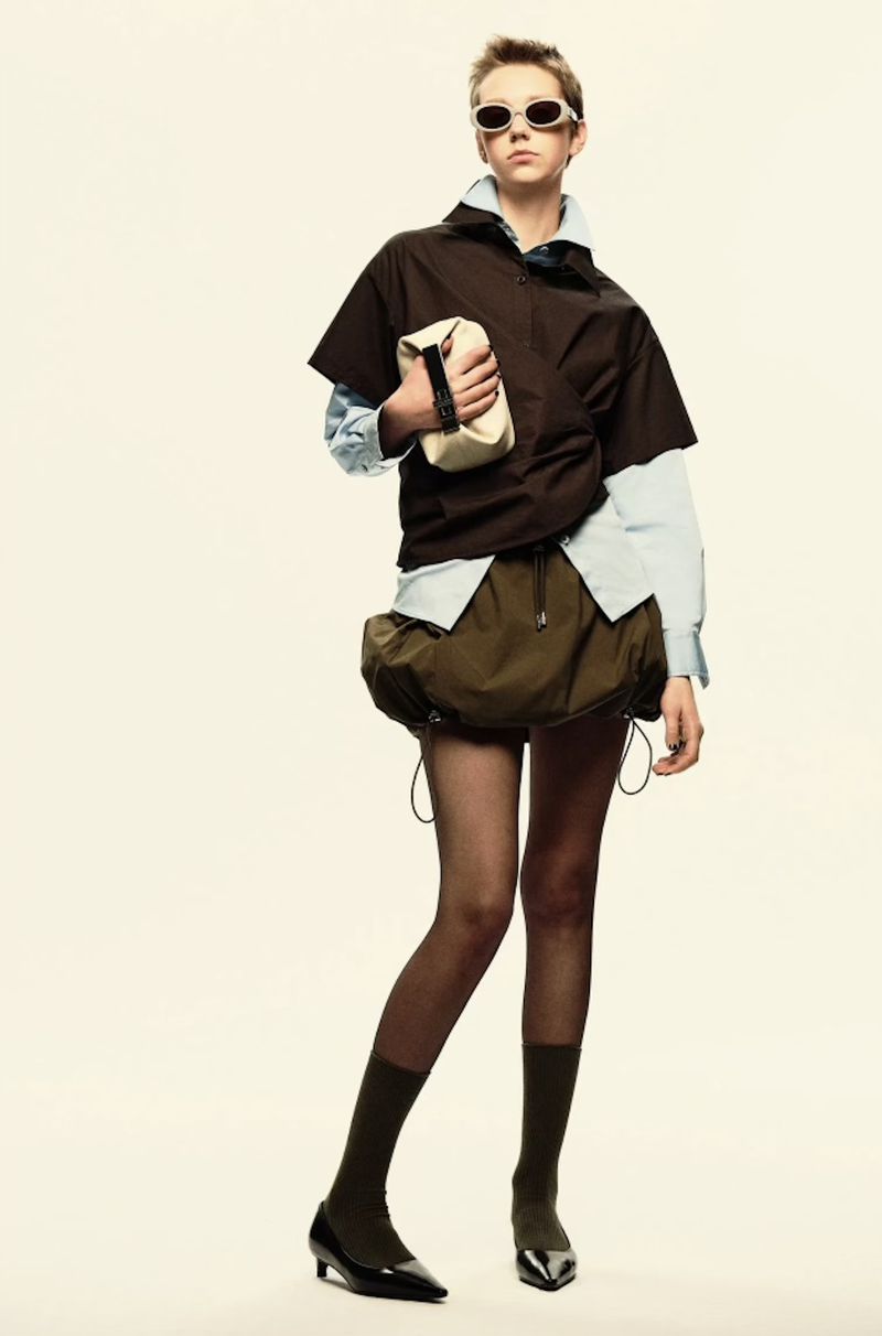 A fashionable young adult poses in a studio wearing a layered brown and light-blue shirt, olive skirt, dark tights, and holding a beige bag.