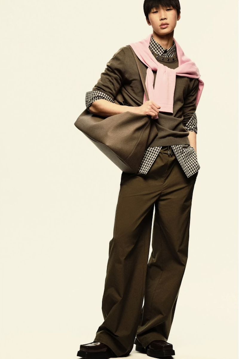 An Asian male model poses in a studio wearing an olive-green outfit and loafers, carrying a large tote bag with a pink sweater around his neck.
