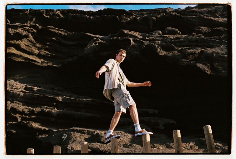 Young man walks across wooden posts near a dark rock formation. Wearing casual clothing, he balances with arms outstretched.