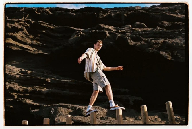 Young man walks across wooden posts near a dark rock formation. Wearing casual clothing, he balances with arms outstretched.