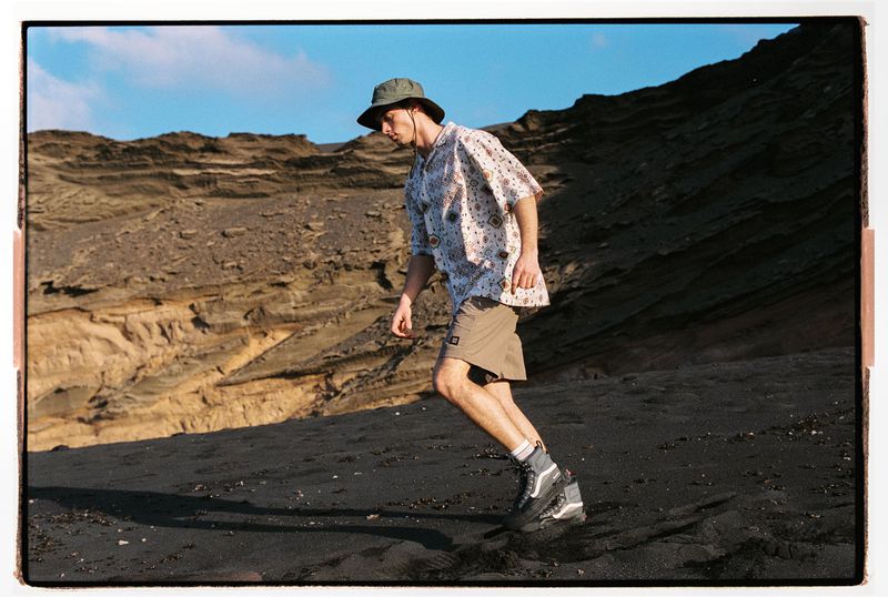 Man walking on black sand beach, wearing hat, patterned shirt, and hiking boots, with layered cliffs and blue sky background.