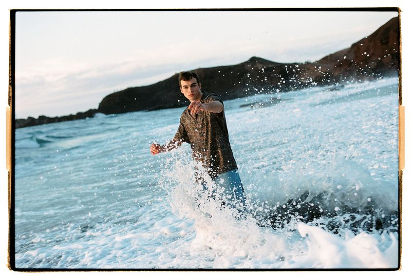 A young man stands in ocean waves on a rocky beach, pointing at the viewer, with a mountain in the background under an evening sky.
