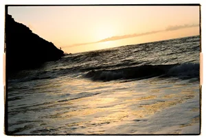 Ocean waves crashing against a rocky coast at sunset with a person standing on the cliff edge, captured on film.