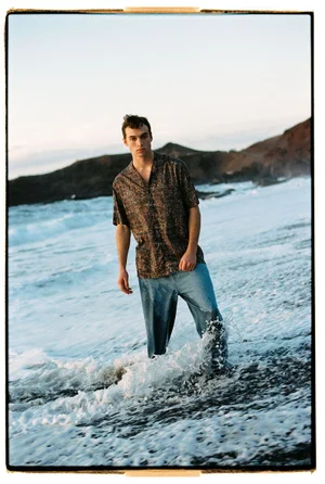 A man wearing a patterned short-sleeve shirt and denim jeans stands in crashing ocean surf on a coastal beach.