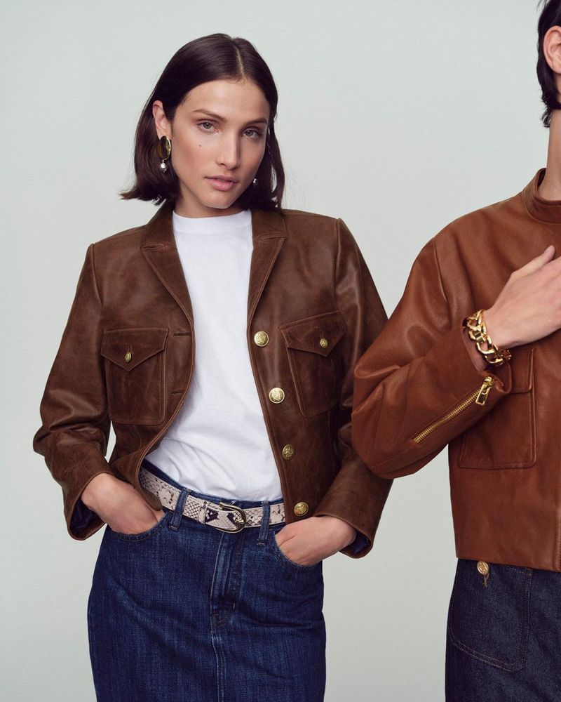 Two fashion models in brown leather jackets, white shirts, and denim skirts. One model faces the camera, while the other is partially visible.