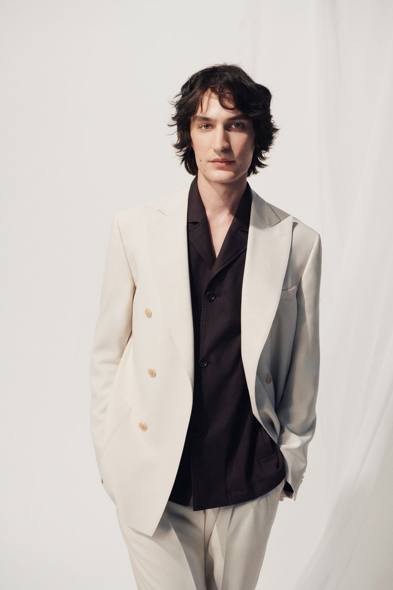 Portrait of a brunette man in a light beige suit jacket and matching trousers against a plain white backdrop in a studio setting.