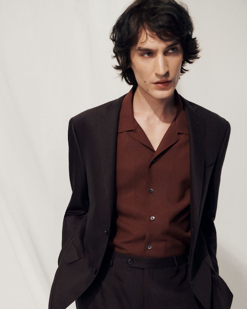 Portrait of a male fashion model in a dark brown suit and open-collar shirt, looking off-camera against a neutral studio backdrop.
