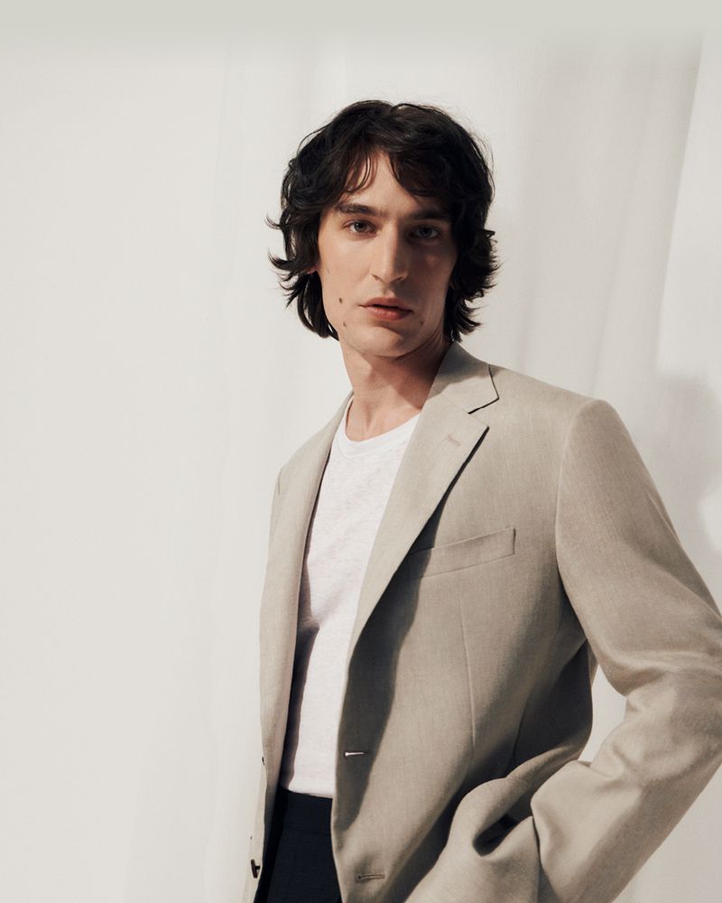 Portrait of adult male with dark wavy hair, wearing a white t-shirt and light beige blazer against a light neutral background in studio lighting.