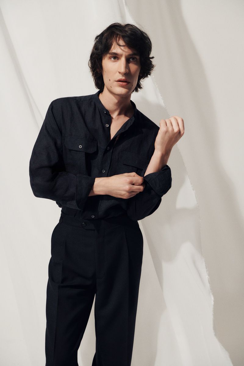 Studio shot of an adult male fashion model in a dark, collared shirt rolling up his sleeves while posing in front of a white backdrop.