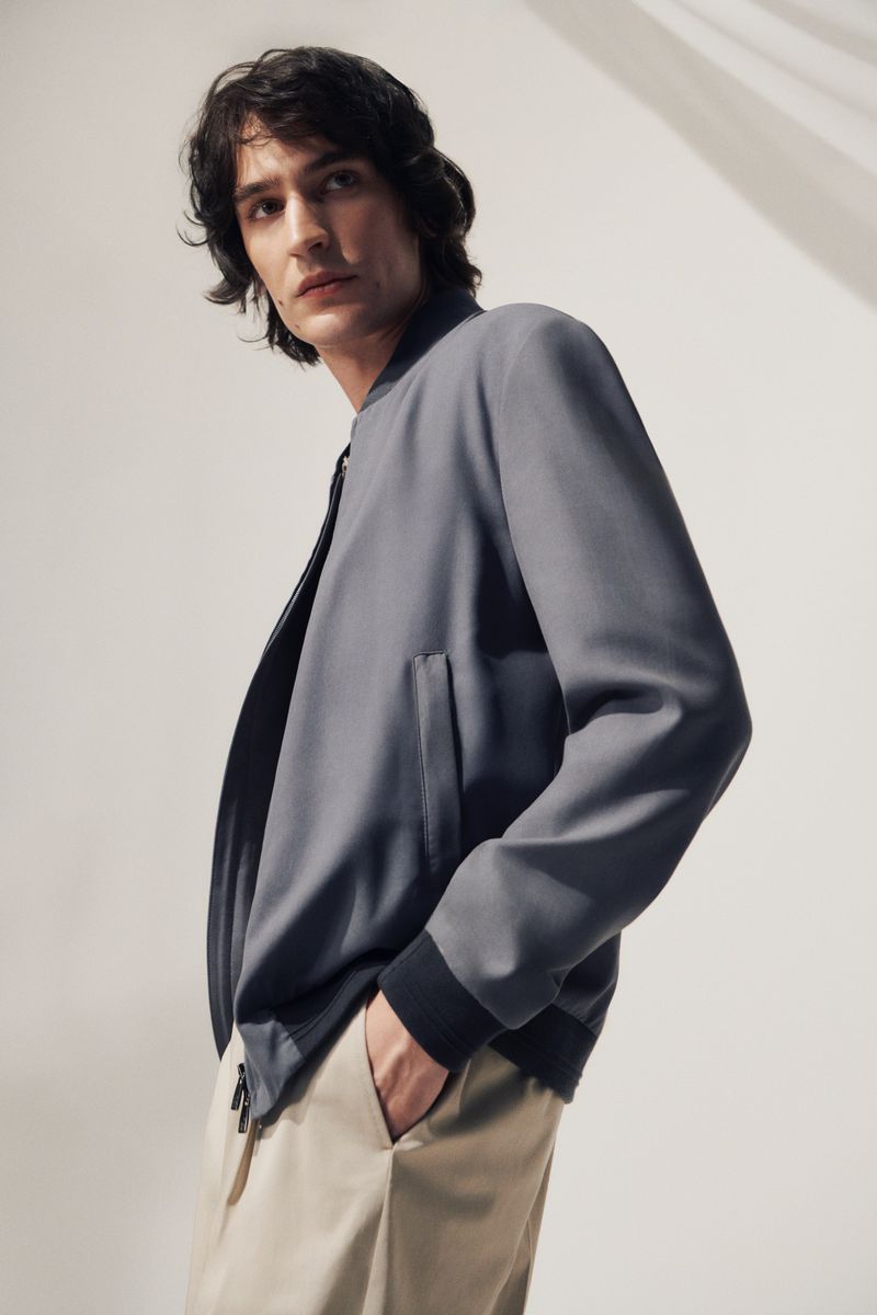 Man in a gray bomber jacket and beige pants posing in a studio with soft, diffused lighting showcasing a modern, fashionable look.
