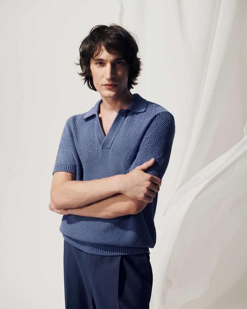 A stylish adult man in a blue knit polo shirt and dress pants stands in a photo studio against a white backdrop, fashion portrait.