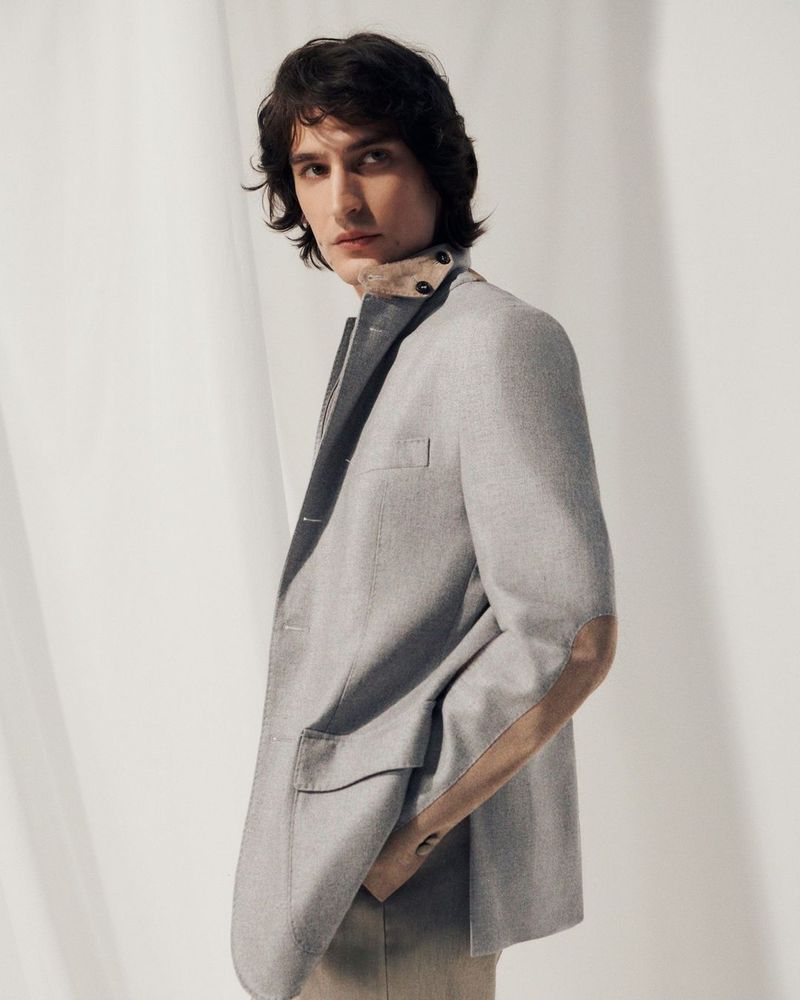 Model wearing a grey blazer jacket against a pale draped backdrop for Corneliani SS24.