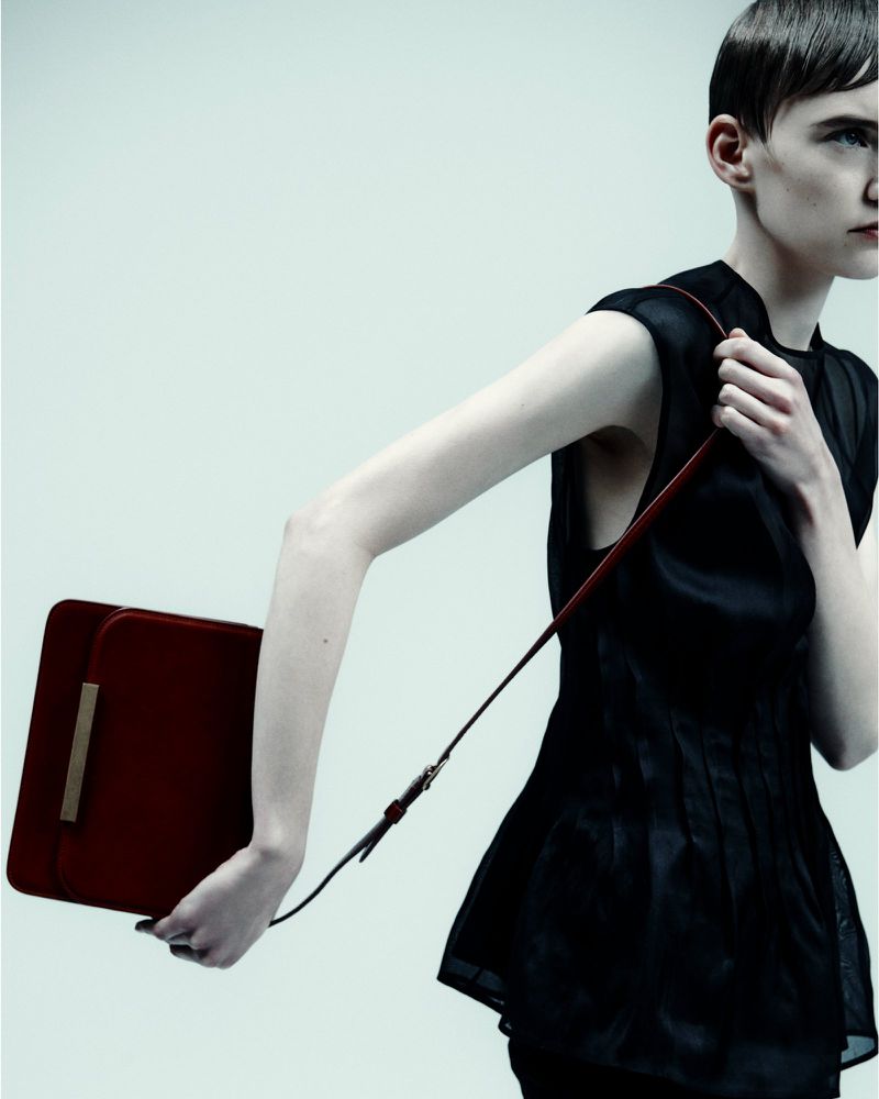 A fashionable adult female model poses in a studio with a red handbag. She is wearing a dark, sleeveless blouse and has short hair.