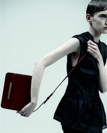 A fashionable adult female model poses in a studio with a red handbag. She is wearing a dark, sleeveless blouse and has short hair.
