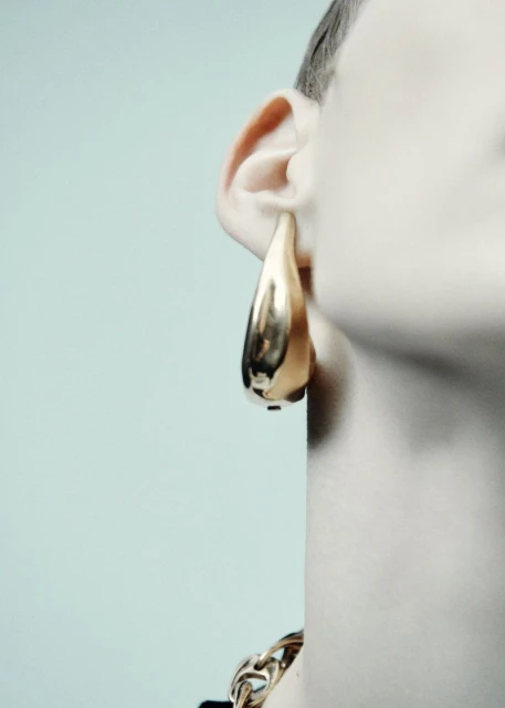 Close-up of a model's ear with a gold, tear-drop shaped earring and a gold chain necklace against a light blue background.