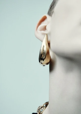 Close-up of a model wearing a large sculptural gold teardrop earring and chunky gold chain against a pale blue backdrop.