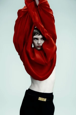 Fashionable woman with short hair poses in studio. She wears black trousers with a gold belt and is partially covered by red silk fabric.