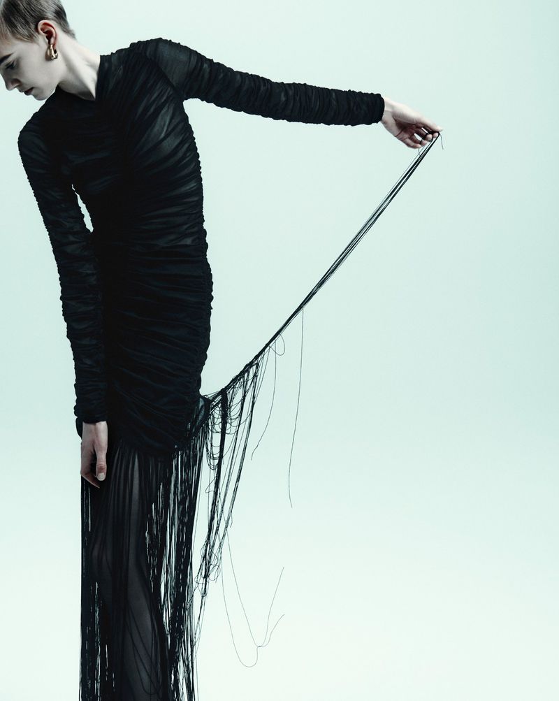 A slender person in a dark, draped gown holds loose threads against a bright backdrop, emphasizing texture and unique silhouette in a fashion setting.