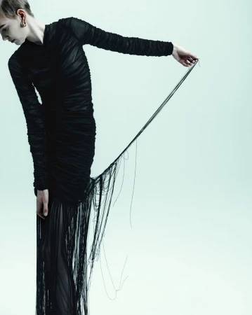 A slender person in a dark, draped gown holds loose threads against a bright backdrop, emphasizing texture and unique silhouette in a fashion setting.