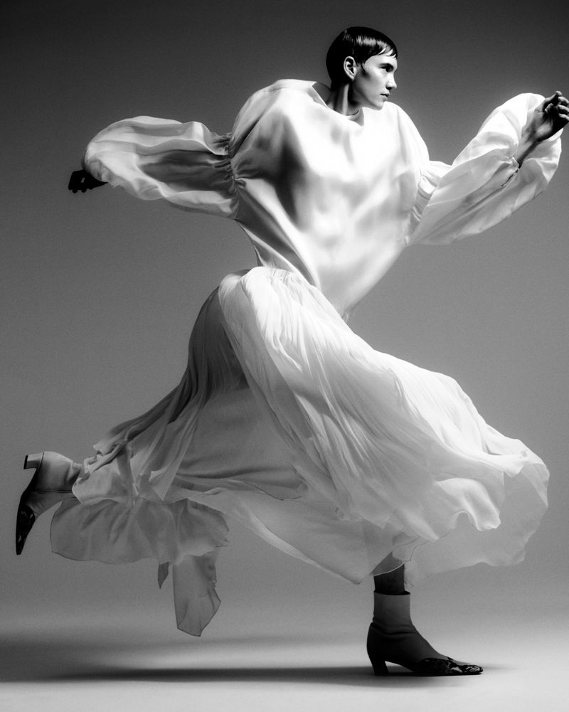 Monochrome fashion shot of a model in mid-air wearing a long, flowing white dress with dramatic sleeves, paired with heeled ankle boots.