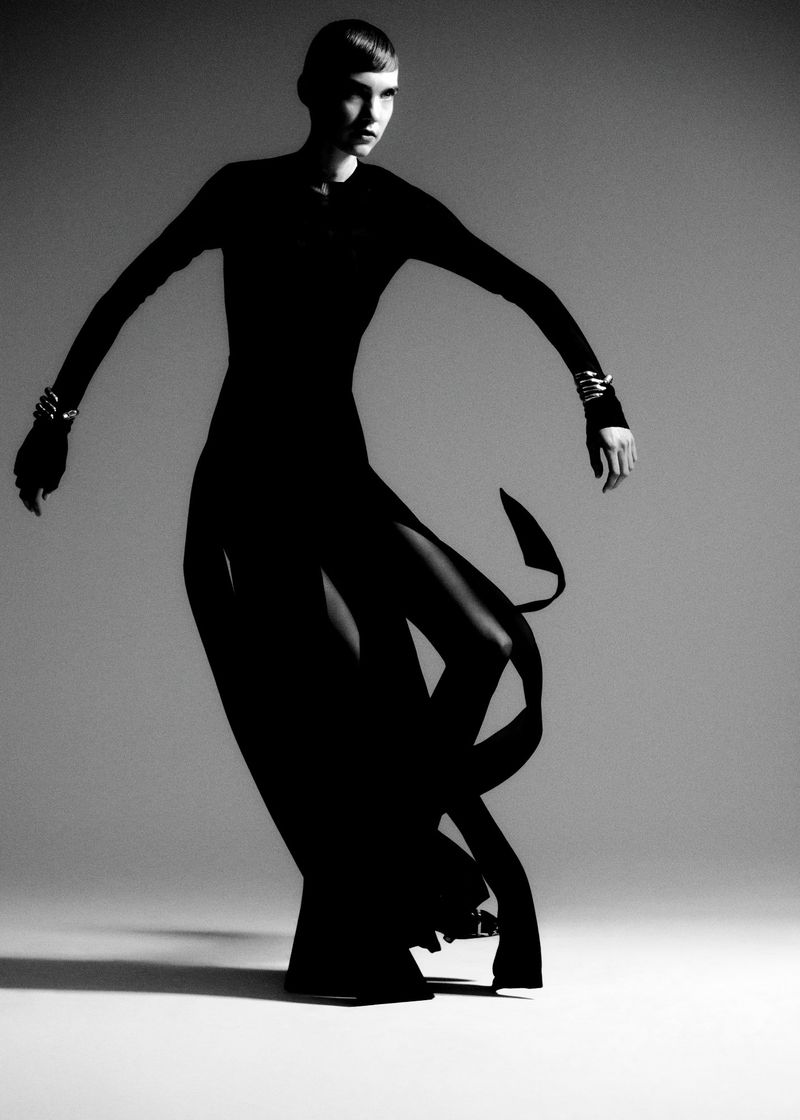Elegant woman in a black dress and metal bracelets poses in a studio, showcasing fashion in a high-contrast black and white photograph.