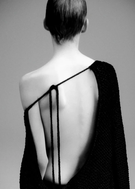 Monochrome shot from the back of an adult female wearing a black knit garment with a single strap over her shoulder in a studio.