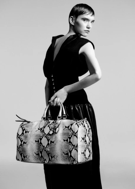 Monochrome portrait of a woman in a black dress, looking over her shoulder while holding a snakeskin pattern bag in a studio.
