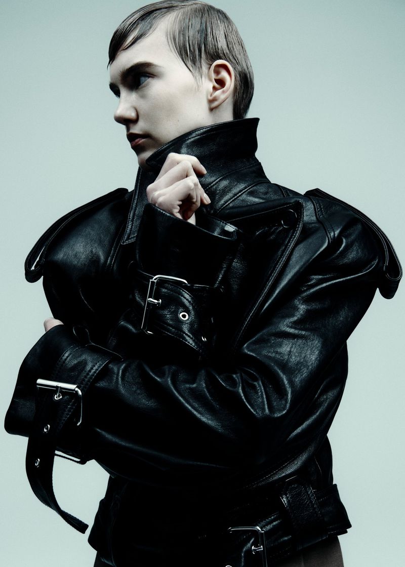 Adult poses in a dark leather jacket against a light background, showcasing fashion and style in a studio setting.