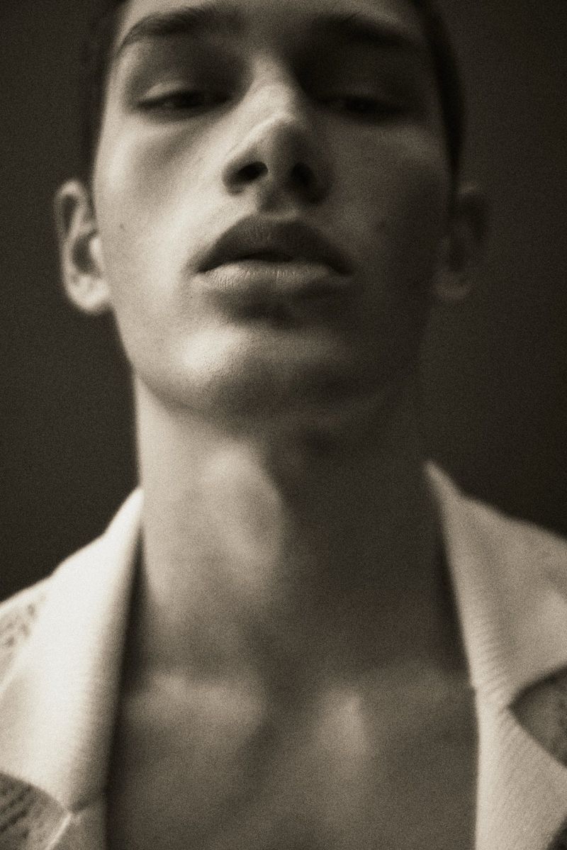 Close-up studio portrait of a young adult male with an open collared shirt, focusing on his face and upper chest in neutral lighting.