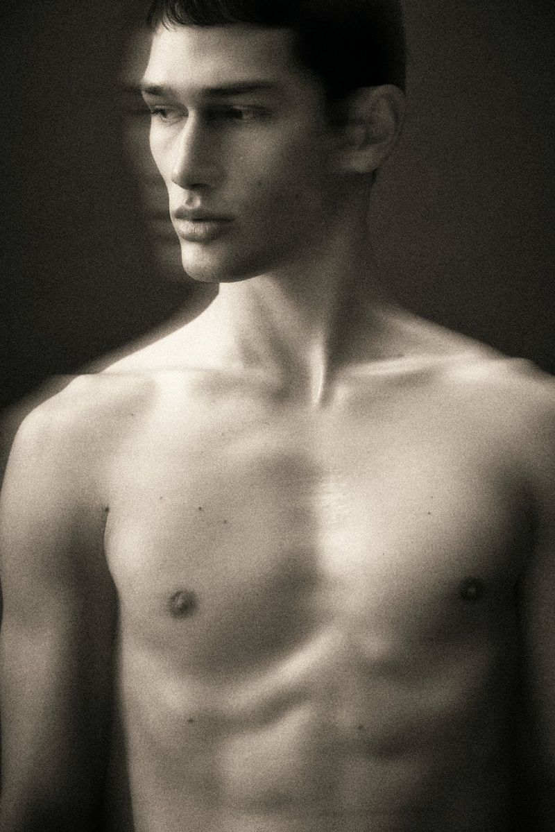 Portrait of an adult male with short dark hair, standing topless in front of a blurred background with soft, neutral lighting.