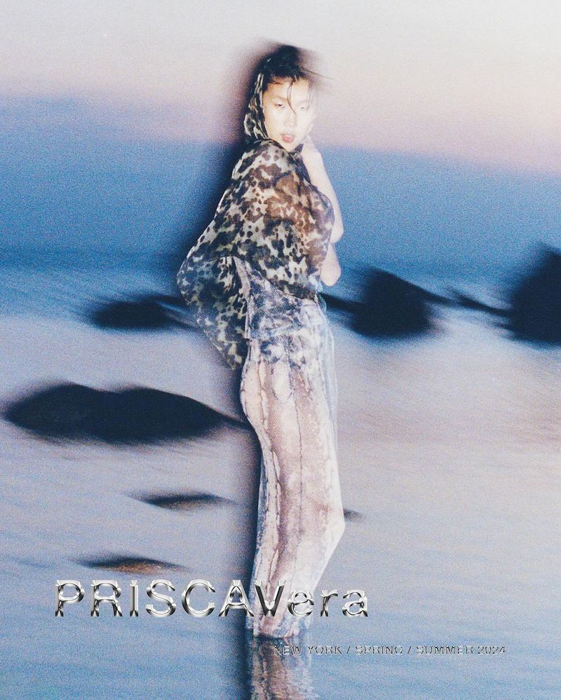 A woman stands in water on the beach wearing a transparent, patterned hooded top and pants, with a soft sunset in the background.