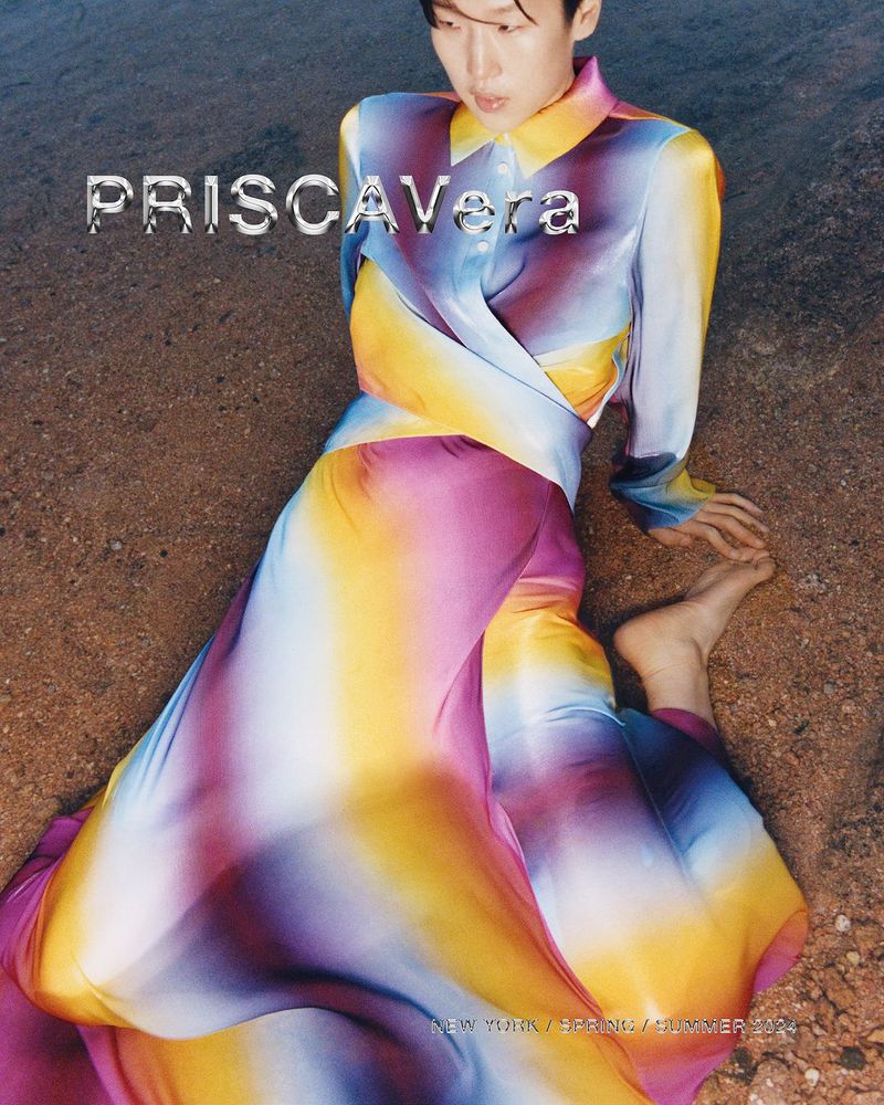 Editorial shot of a person in a colorful silk dress, lying on sand with a glossy magazine aesthetic. The dress features blue, pink, and yellow tones.
