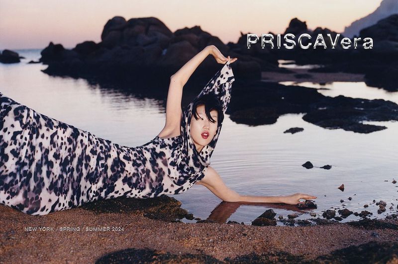 A woman in a printed dress lies on a beach at sunset, posing for a fashion editorial. She has short dark hair and is facing the camera.