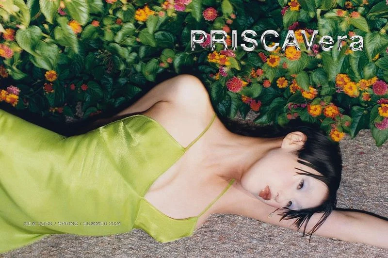Woman in a light green dress lying on the ground next to vibrant flowers. Fashion editorial image for Priscavera New York collection.