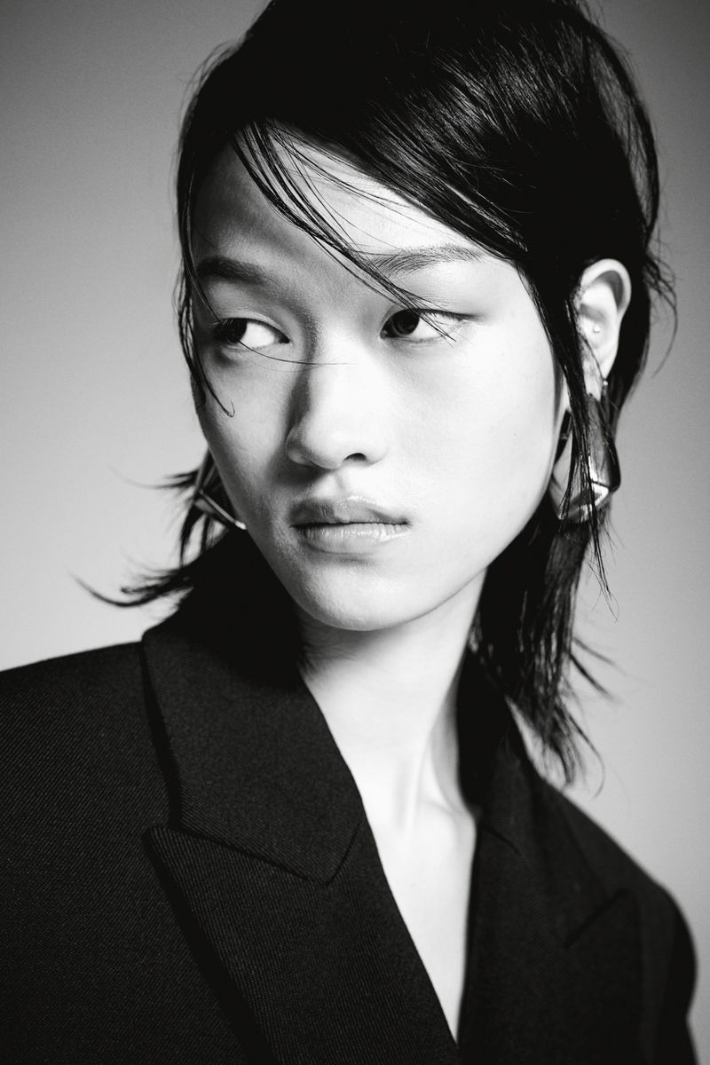 Monochrome portrait of an Asian female model with dark hair wearing a black blazer and geometric earrings against a grey backdrop.