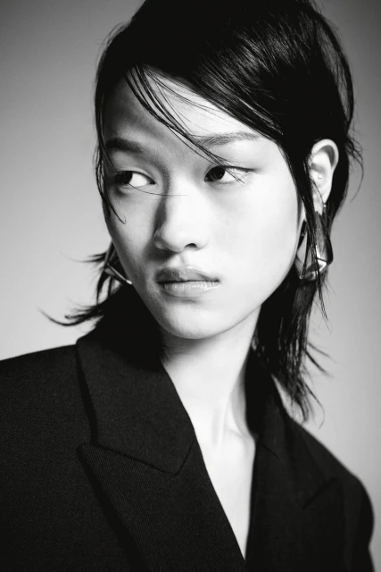 Monochrome portrait of an Asian female model with dark hair wearing a black blazer and geometric earrings against a grey backdrop.